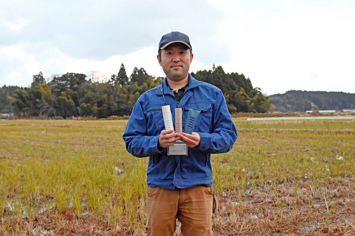 有機農業に取り組む田んぼを背景に、NAIAの新商品を手にする脊戸さん